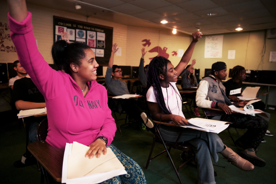 Picture of a classroom, two teenage girls raising their hand at their desks